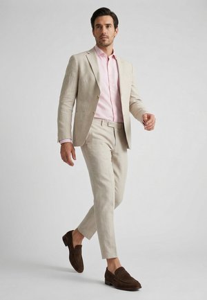 Man loopt, draagt een beige pak met een lichtroze overhemd en donkerbruine loafers tegen een effen lichte achtergrond.