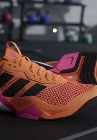 Chaussures de sport orange et rose avec des bandes noires Adidas, exposées sur une surface sombre dans une salle de sport intérieure.