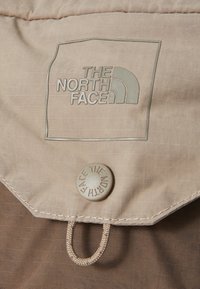 Beige szövet, textúrált mintázattal, kiemelt logóval, amelyen a "THE NORTH FACE" felirat szerepel, négyzet alakú foltban, valamint egy kerek gomb a legalján.