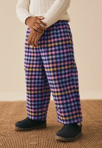 Rutet bukser i lilla, pink og gul, lavet af et blødt materiale, med brede ben. Sorte støvler med gummisål og lynlås på siden.