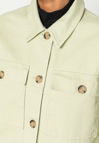 Světle zelená denimová bunda s límcem, dvěma náprsními kapsami a hnědými knoflíky. Má texturovanou tkaninu a viditelné švy.