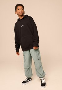 Schwarzer Kapuzenpullover mit Vordertasche, kombiniert mit hellgrünen Cargo-Hosen mit Seitentaschen und schwarzen Sneakers mit weißen Akzenten.