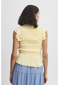 Top amarillo con fruncidos y mangas acampanadas, con un dobladillo tipo peplum, que presenta una tela texturizada. Usado sobre una falda de mezclilla azul.