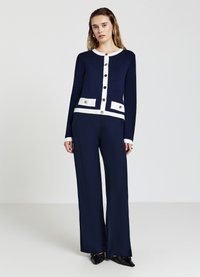 Cardigan blu navy con accenti bianchi e bottoni dorati, abbinato a pantaloni ampi coordinati. Tessuto liscio, stile aderente.