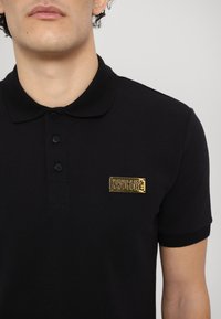 Črna polo majica iz bombažne mešanice, z ovratnikom in ploskim zapiranjem na tri gumbe, okrašena z zlatim logom na prsih.
