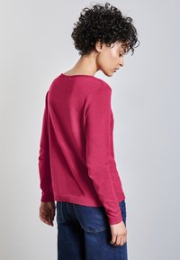 Magenta gestrickter Pullover mit Rundhalsausschnitt, langen Ärmeln, gerippten Bündchen und Saum, kombiniert mit dunkelblauen Jeans.