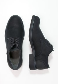 Chaussures noires en tissu texturé, bout rond, lacées et avec une semelle en caoutchouc. Une chaussure a un petit talon ; les deux présentent un design minimaliste.