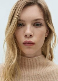 Zbliżenie na osobę z falującymi, jasnoblond włosami, ubraną w prążkowany, beżowy sweter dzianinowy. Skóra jest lekko opalona, a makeup naturalny.