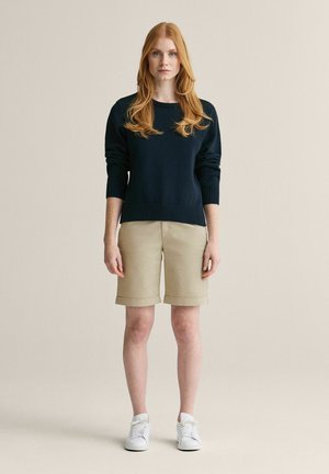 Kvinde med langt rødt hår iført en sort sweater, beige knælange shorts og hvide sneakers, står mod en ensfarvet beige baggrund.