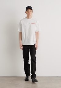Hvit bomull t-skjorte med rødt "Calvin Klein"-logo, kombinert med sorte jeans og svarte sko. Modellen har på seg en svart cap og står mot en ensfarget bakgrunn.