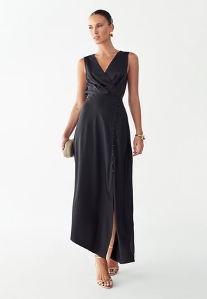 Vrouw draagt een zwart, mouwloos wikkeljurkje met zijknopen, houdt een beige clutch vast en draagt gouden oorbellen, een armband en sandalen met bandjes.