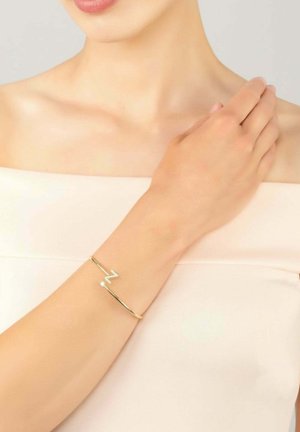 Gold cuff bracelet featuring a stylized "Z" accent with a small crystal detail. Smooth metal surface, minimalist design, worn on a wrist.
