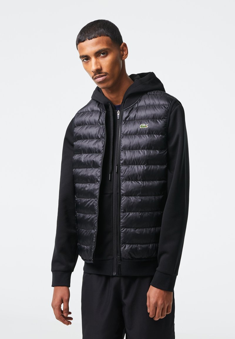 Gilet à capuche noir en matelassé, avec zip et logo Lacoste. Superposé à un sweat à capuche noir.