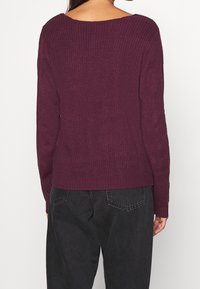 Burgundy stickad tröja med vid halsringning, ribbad textur, långa ärmar och kortare nederkant, kombinerad med svarta högmidjade byxor.