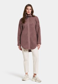Fleece-pullover i dæmpet mauve med høj krave, frontlynlås og forlænget søm, parret med hvide bukser og lyse sneakers.