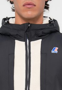 Must mustaga pinda jakk, millel on valge fleece-vooder, kapuuts, millel on reguleeritav nöör, ja K-Way logo vasakul rinnal. Ees lukuga avamine ja taskud.