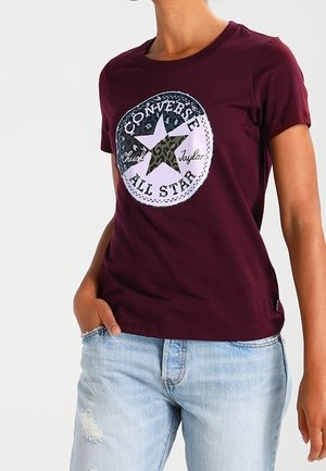 Kobieta ubrana w bordową koszulkę Converse All Star z logo gwiazdy w kamuflażu, połączoną z jasnoniebieskimi podartymi jeansami, ręka w kieszeni.