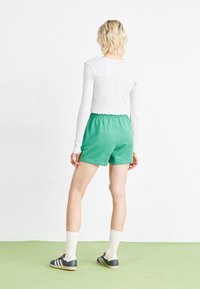 Bílý top s dlouhým rukávem a žebrovanou texturou, doplněný zelenými bavlněnými šortkami s elastickým pasem. Černé boty s bílými pruhy a ponožky po kolena.