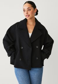 Schwarzer doppelreihiger Mantel mit großen Revers, weiten Ärmeln und Seitentaschen. Auffälliger Textilstoff mit Knopfakzenten.