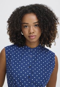 Mladá žena s kučeravými vlasmi, oblečená v tmavomodrej blúzke bez rukávov s bodkami, stojaca čelom k nám s neutrálnym výrazom.