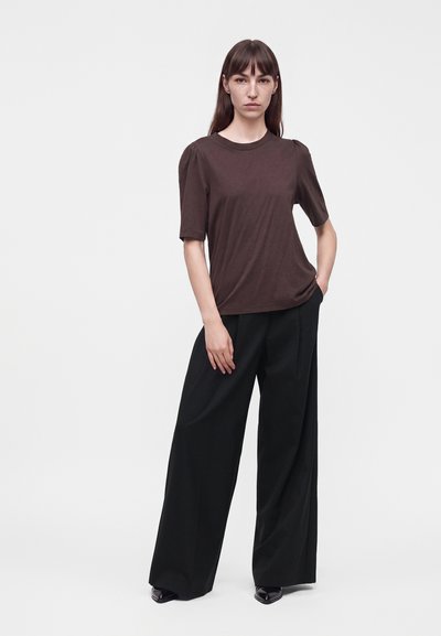 Femme aux longs cheveux marron portant un haut marron à manches courtes et un pantalon noir à jambes larges, debout avec une main dans la poche.