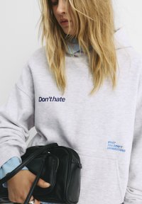 Felpa grigia chiara con testo ricamato blu navy e blu. Presenta una tasca frontale ed è sovrapposta a una camicia a quadri blu. Borsa nera in mano.