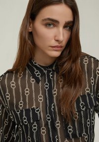 Camicia nera trasparente con motivo a catena. Presenta due tasche sul petto e un colletto classico. Texture morbida e design leggero.