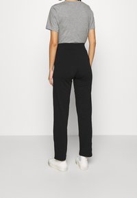 Schwarze, locker sitzende Hose mit hohem Bund, kombiniert mit einem grauen T-Shirt. Das Model steht vor einem neutralen Hintergrund und trägt weiße Schuhe.