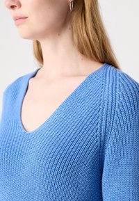 Femme portant un pull en maille bleu clair à col en V, montrant les détails de l'épaule et du cou, avec une boucle d'oreille argentée en forme d'anneau à une oreille.
