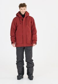 Rote wasserdichte Jacke mit hohem Kragen und Fronttaschen, kombiniert mit schwarzen wasserdichten Hosen und Skistiefeln. Glatte Textur, praktisches Design.