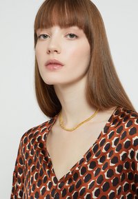 Ruskea ja kerma leopardikuosiset paita syvällä V-pääntiellä. Keltainen helmikaulakoru, sileä tekstuuri, jota mallilla on päällään pitkät, suorat hiukset.