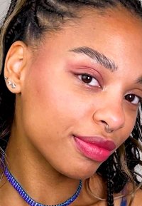 Primer plano de una cara con piel marrón clara, cabello trenzado, anillo de nariz de aro plateado y pendientes de perno plateados. Los labios tienen un degradado de tonos rosados.