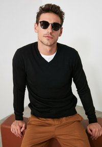 Pull noir à col en V au design ajusté, associé à un pantalon marron. L'homme porte également des lunettes de soleil rondes, assis sur un ottoman en simili cuir marron.