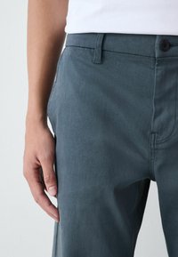 Eine Person trägt eine dunkelgraue Hose und ein weißes Hemd, mit der linken Hand entspannt an der Seite vor einem schlichten Hintergrund.