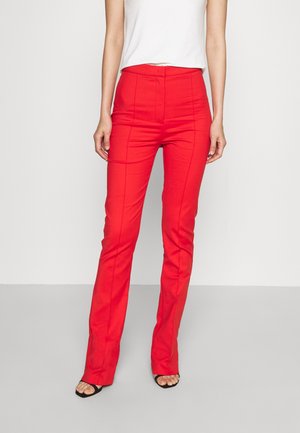 Femme portant un pantalon ajusté rouge vif taille haute avec des coutures devant, associé à un haut blanc sans manches et des talons ouverts noirs.