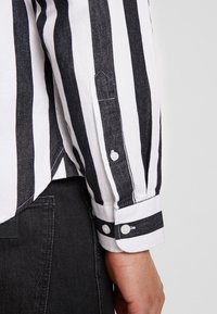 Zwart-wit gestreept shirt met een close-up van een knoopmanchet, met een textuurstof en contrasterende knoopdetails.