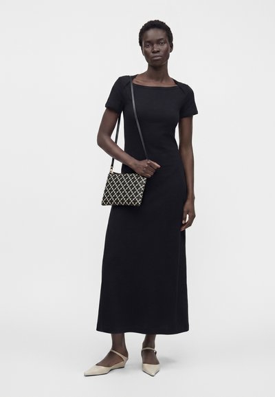 Femme portant une robe longue noire à manches courtes, des ballerines beige pointues et un sac bandoulière à motif noir et blanc sur un fond clair uni.
