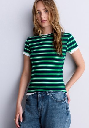 Jeune femme aux longs cheveux blonds portant un t-shirt rayé vert et bleu marine et un jean bleu sur un fond uni.