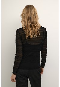 Svart långärmad topp med texturerat genomskinligt tyg, med horisontella ränder. Bärs med randiga byxor, sedd bakifrån.