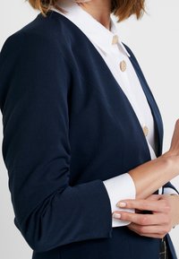 Námořnický přizpůsobený blazer s dlouhými rukávy, texturovanou tkaninou a bílou košilí pod ním, která má kulaté zlaté knoflíky a strukturovaný límec.