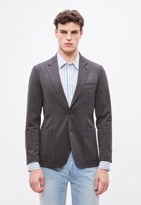 Γκρι σακάκι από υφή υφάσματος, με δύο μπροστινές τσέπες, κλείσιμο με ένα κουμπί και τσέπη στο στήθος, φορεμένο πάνω από ένα ανοιχτό ριγέ πουκάμισο.