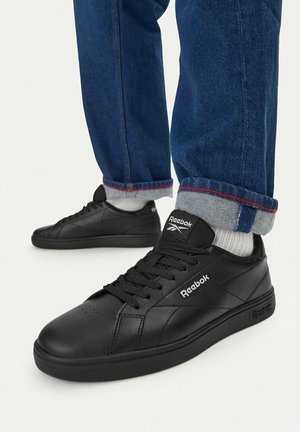 Persona con zapatillas negras Reebok, calcetines blancos y vaqueros azules remangados sobre fondo blanco.