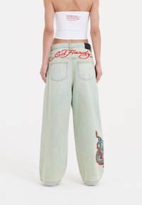 Lichtblauwe wijde jeans met rood geborduurd "Ed Hardy" op de achterkant van de tailleband en ingewikkeld multicultureel borduurwerk op het linkerbeen.