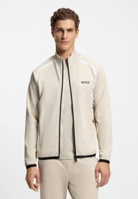 Beige athletic jacket with a high collar, black zipper, and white accents on shoulders. Features a logo on the left chest.