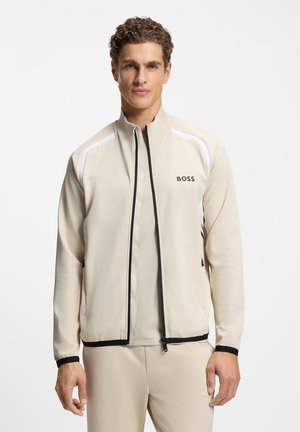 Chaqueta deportiva beige con cuello alto, cremallera negra y detalles blancos en los hombros. Cuenta con un logotipo en el lado izquierdo del pecho.