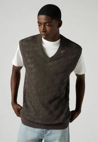 Dunkel gestrickter ärmelloser Pullover mit V-Ausschnitt, der ein strukturiertes Rautenmuster und einen gerippten Saum aufweist, getragen über einem weißen Hemd.
