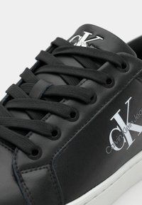 Svart lädersneaker med rund tå, texturerad yta och platta svarta snören. Har en vit "CK"-logotyp och kontrasterande vit sula.