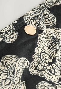 Črna tkanina z belim cvetličnim vzorcem, s kroglo zlatokutonim gumbom, na katerem je vžgano "MINT". Vidne podrobnosti teksturirane površine.