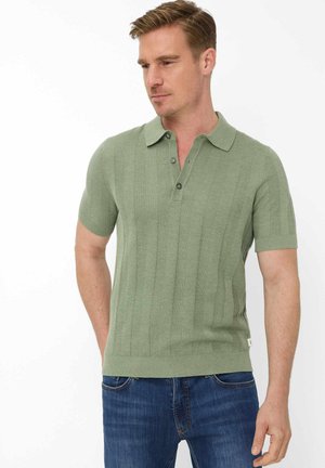 Mann trägt hellgrünes, kurzärmeliges Polohemd mit vertikaler Struktur und blaue Jeans, blickt leicht nach unten und rechts.