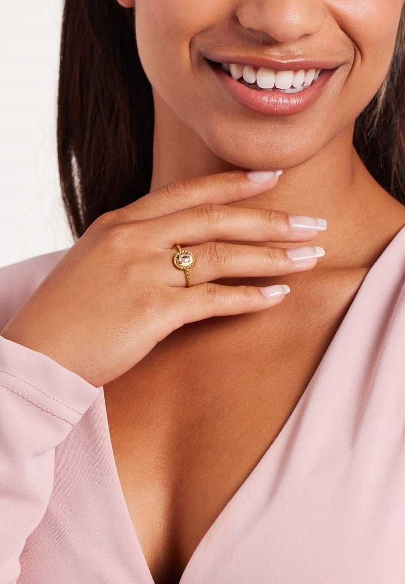Gouden ring met een ovale centrale steen, omringd door kleine accenten. Hand rustend op de kraag van een roze kledingstuk, met natuurlijke nagels.